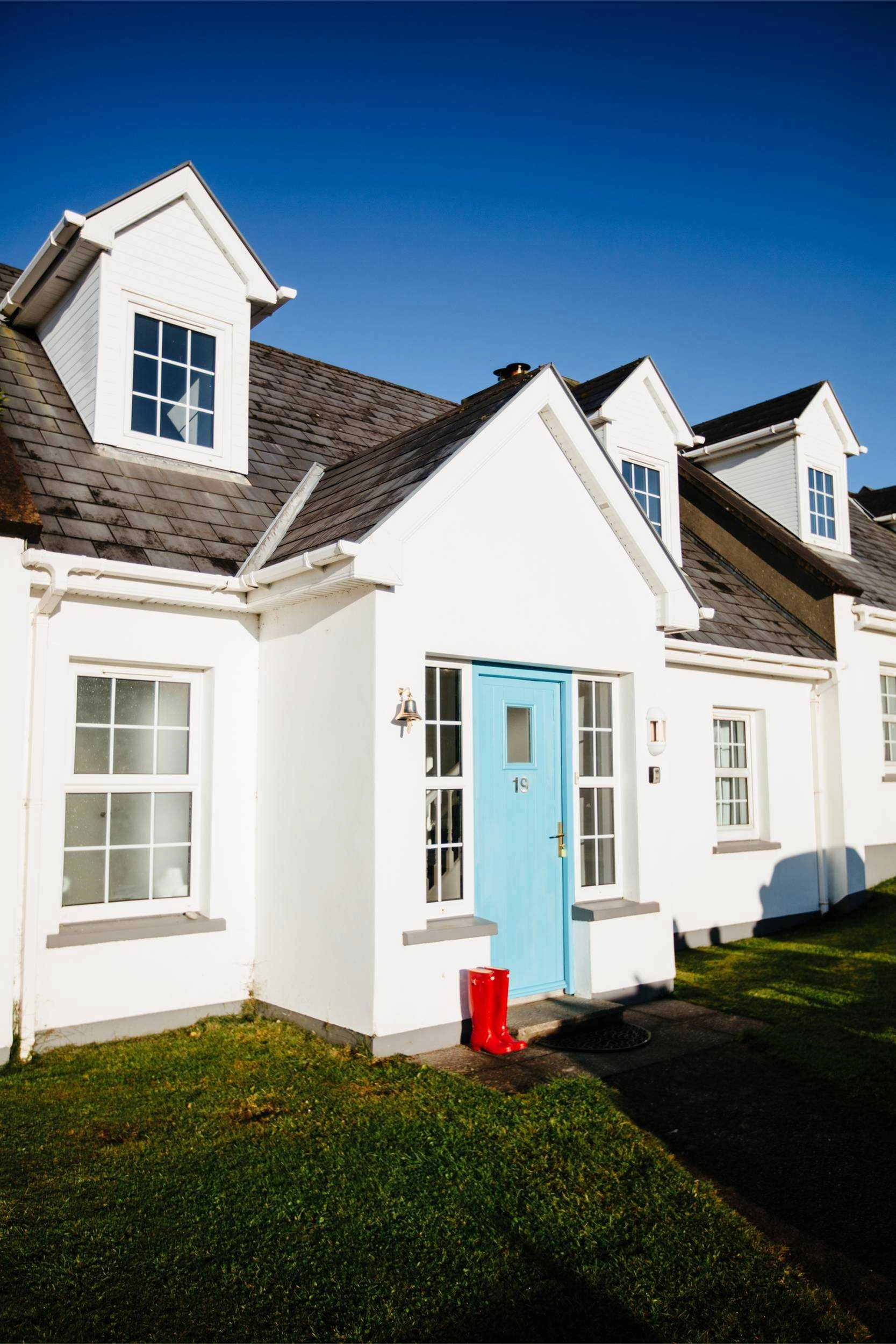 A white cottage with dormer windows and a bright turquoise front door sits on manicured green lawn under clear blue sky. Multiple connected units feature dark slate roofs, white-framed windows, and traditional Irish architectural styling. A red post box stands near the entrance.
