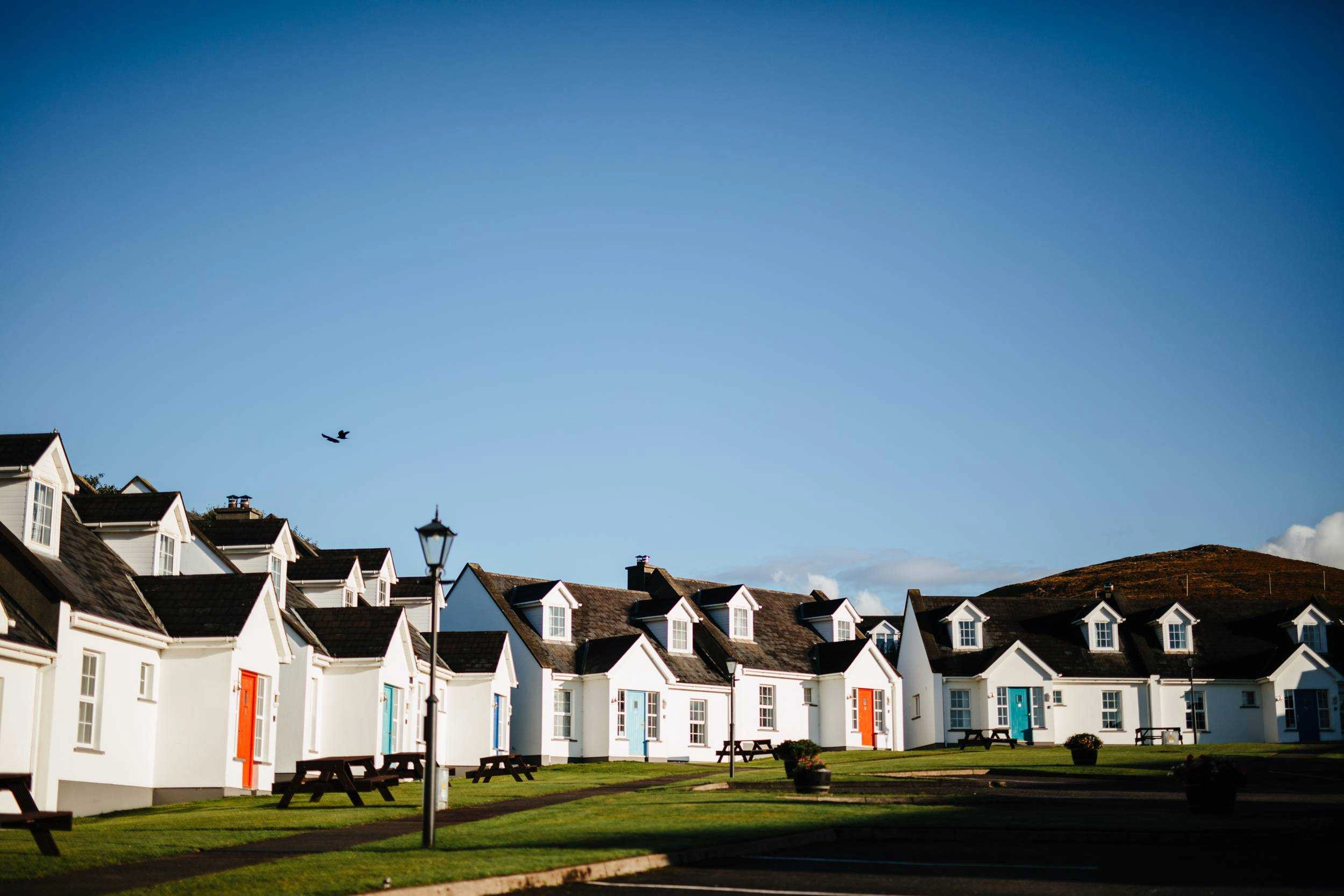 A row of white cottages with dark roofs and dormer windows lined along a manicured green lawn under a clear blue sky, located at Dingle Harbour Cottages.