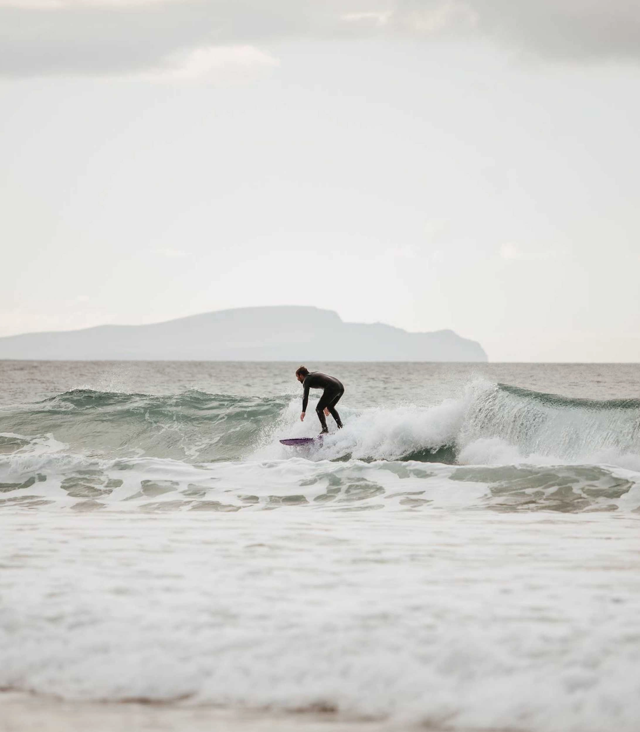 A surfer in a dark wetsuit rides a wave on a white surfboard in the sea, with misty mountains visible on the horizon under an overcast sky.