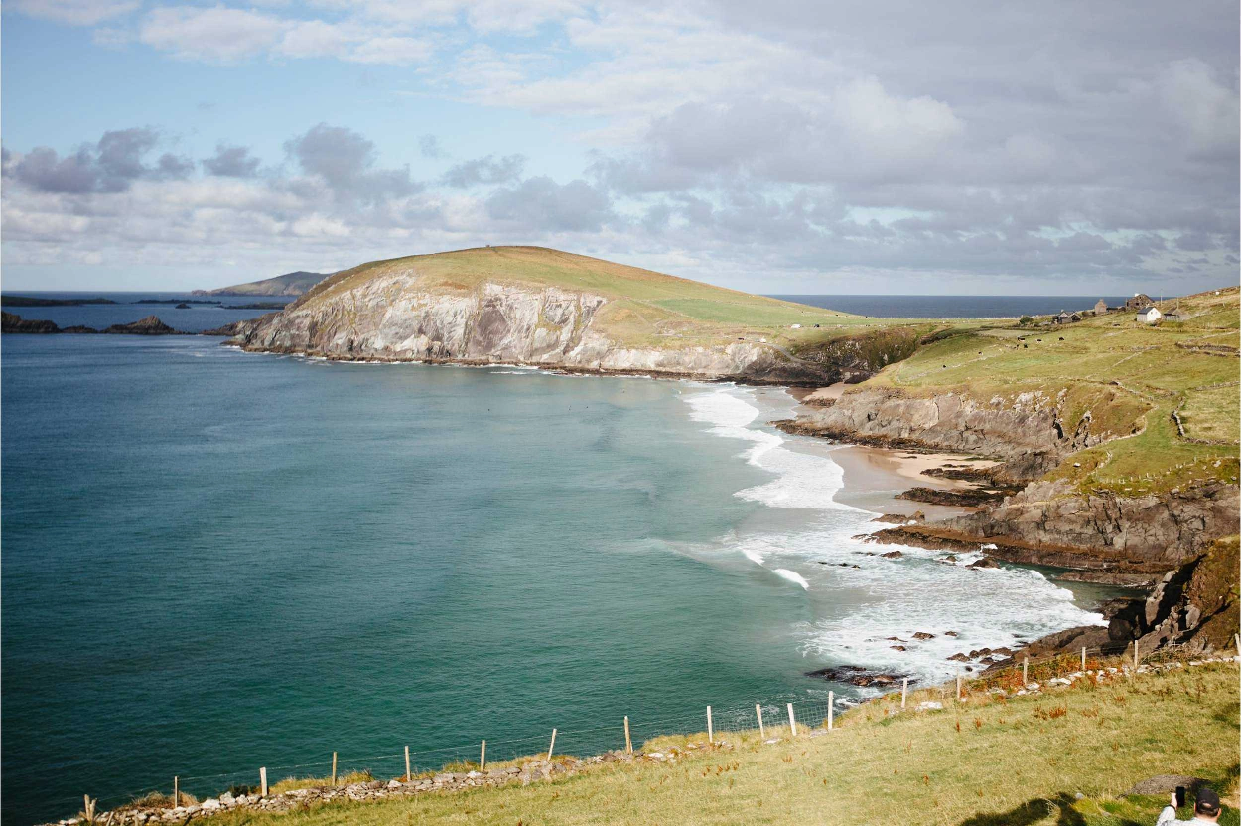Dramatic coastal cliffs with white chalk faces rise above turquoise waters and a sandy beach on the Dingle Peninsula, with rolling green hills and scattered buildings visible on the headland.