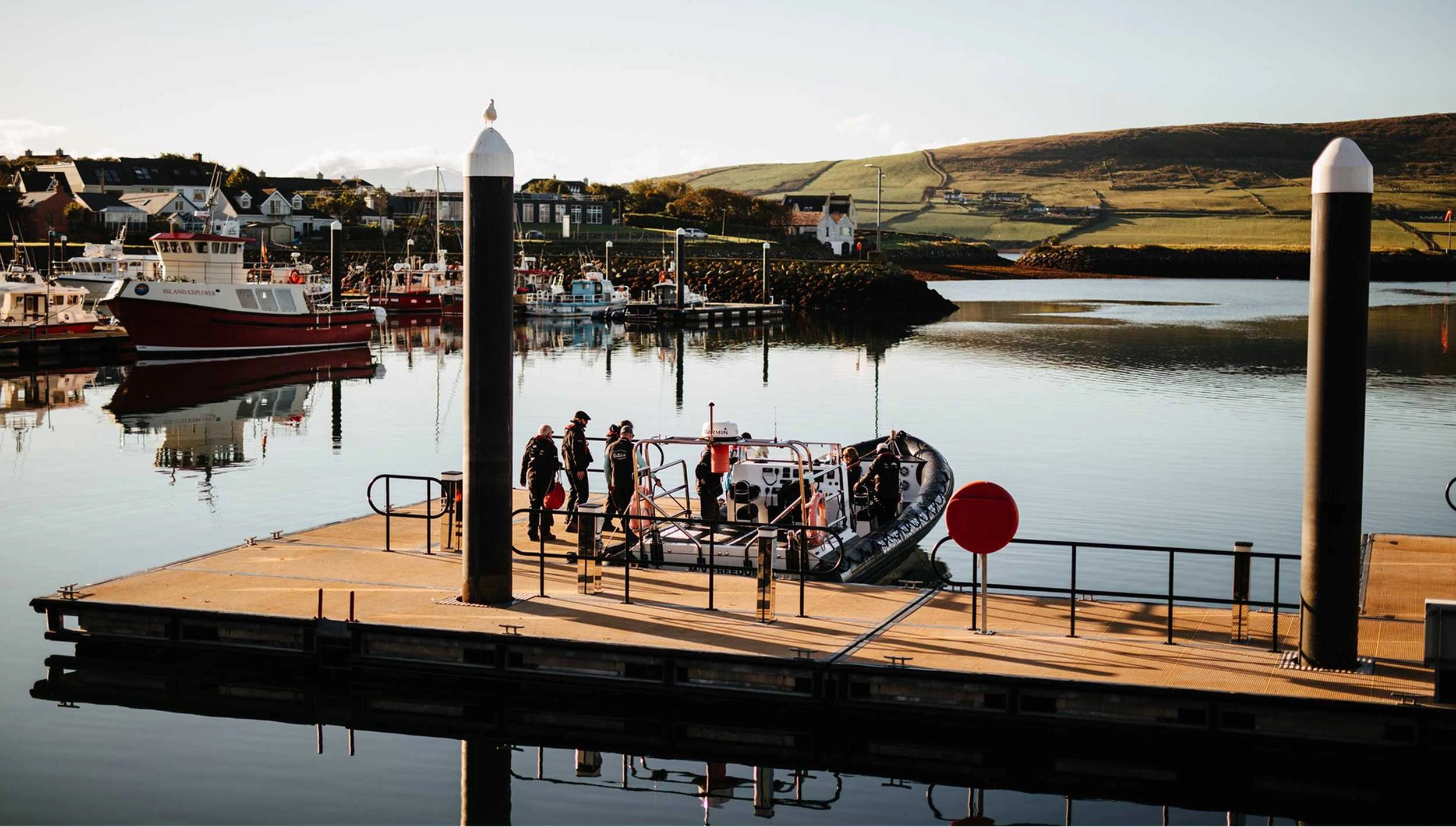 A wooden pier with black railings extends into calm harbor waters, where a group of visitors board a small red and black tour boat. Moored fishing vessels and hillside buildings frame the scenic coastal setting of Dingle Harbour.