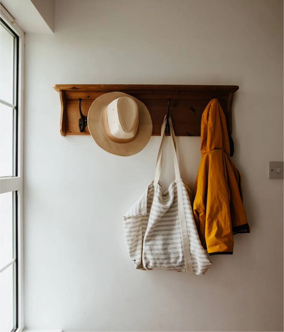 A wooden wall-mounted shelf with hooks holds a tan straw hat, a striped cream and white canvas tote bag, and a mustard yellow folded garment against a white wall next to a bright window.