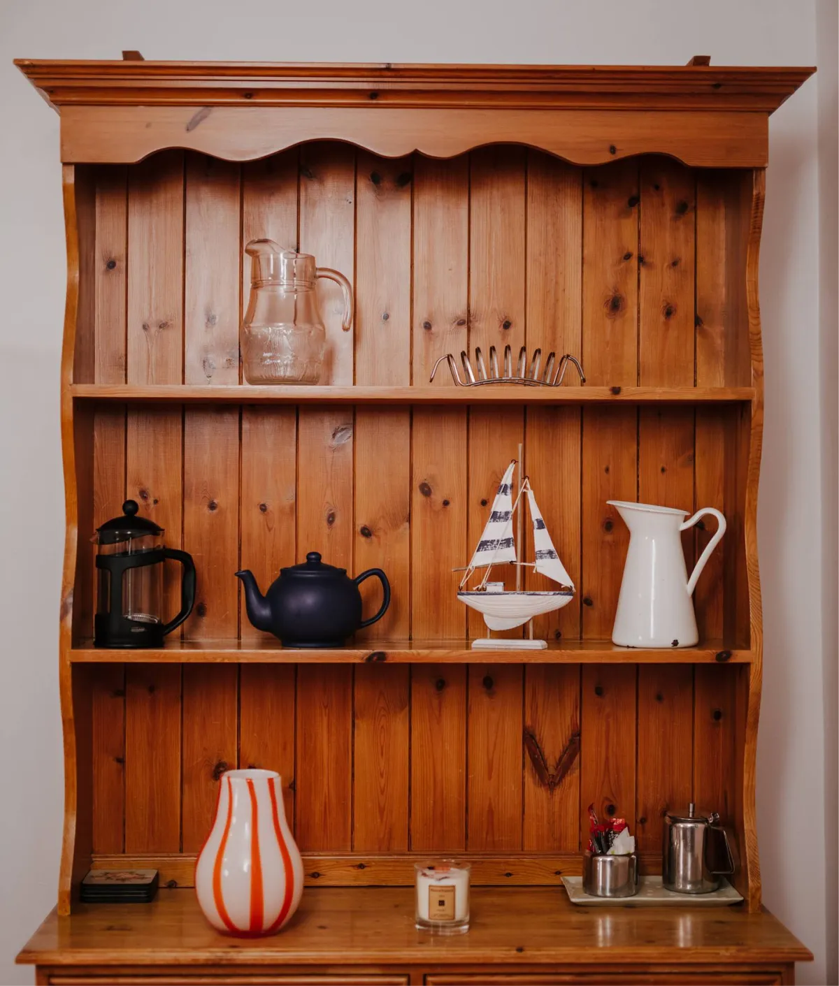 A wooden wall shelf displays nautical and kitchen decor items including black teapots, a white ceramic sailboat, white pitcher, striped orange vase, glass bottles, and decorative accessories arranged across three shelves.