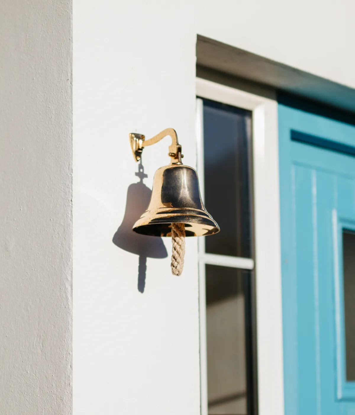 A brass bell mounted on a white wall beside a bright turquoise door, casting a shadow in bright sunlight.