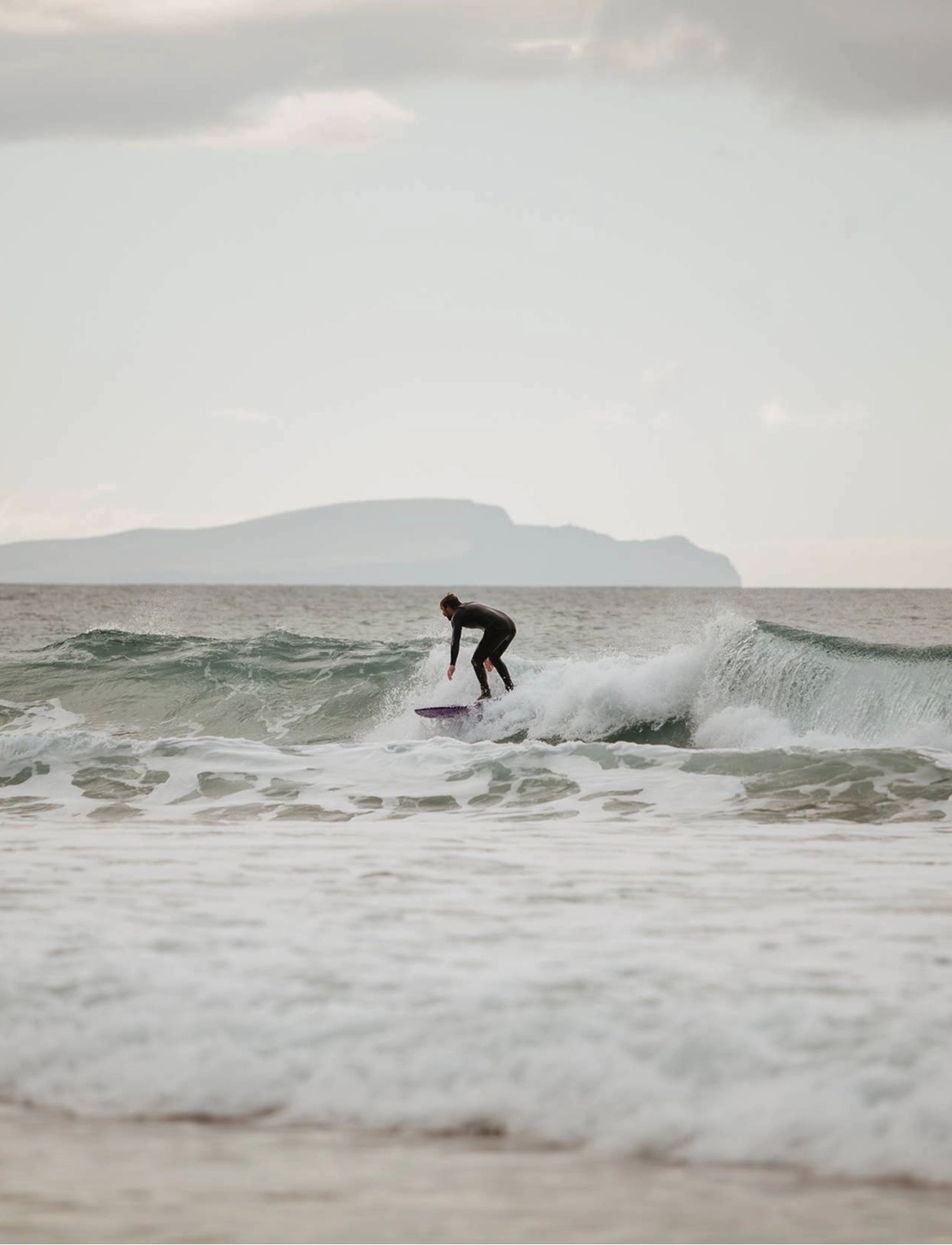 A surfer rides a wave on a beach with a flat-topped mountain visible across the water under an overcast sky.