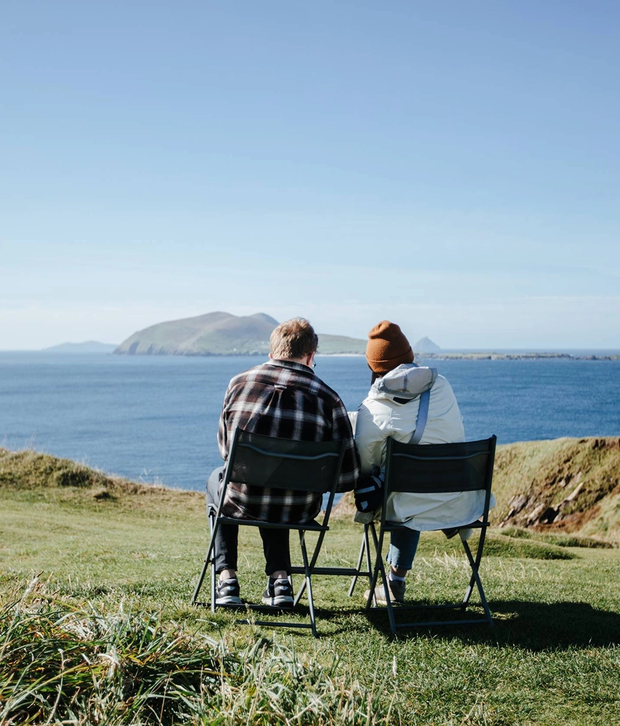 Two people sit in camping chairs on a grassy clifftop overlooking a sparkling blue sea and a distant island under clear skies. They appear to be enjoying a peaceful moment together, gazing out at the scenic coastal landscape.