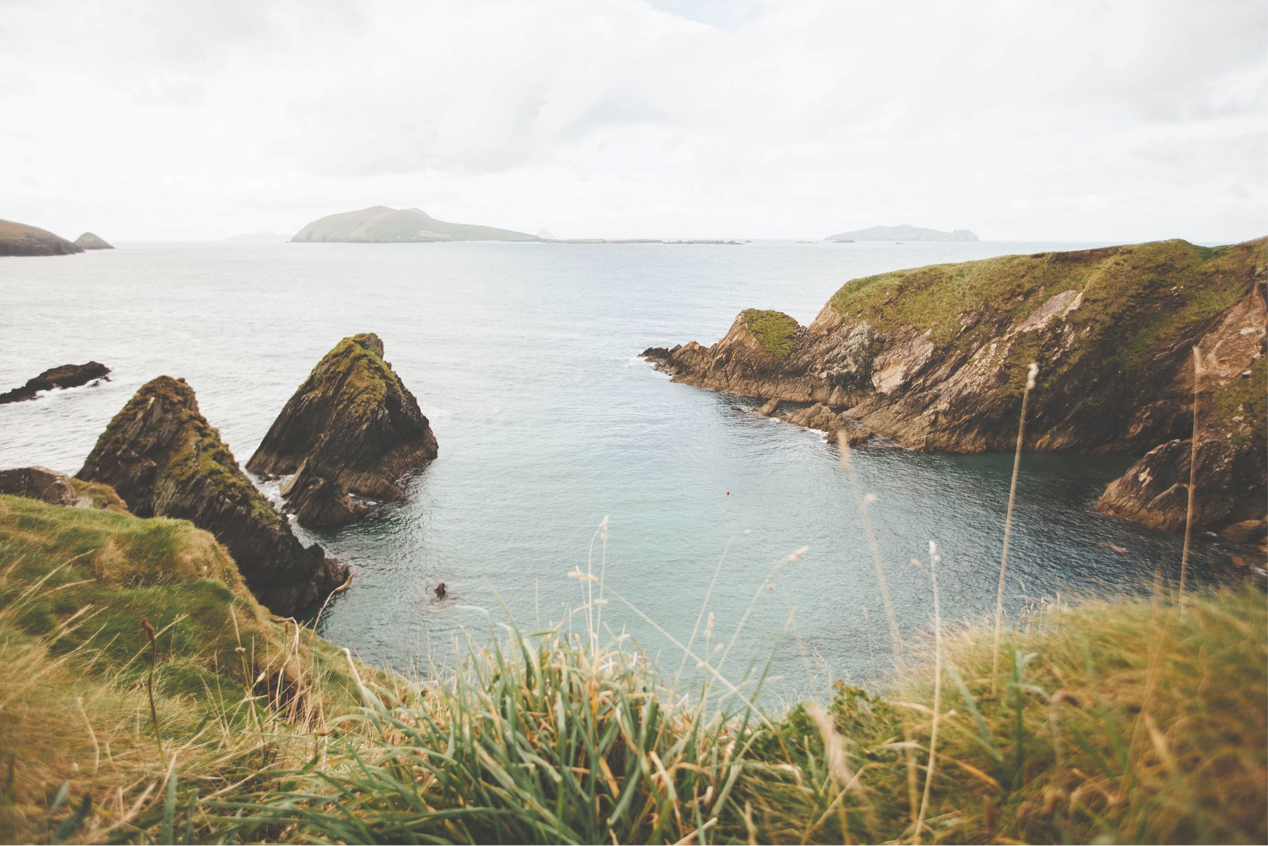 Rugged coastal cliffs and sea stacks overlook a calm turquoise bay with a small sailing boat, surrounded by windswept grasses and rolling headlands along the Dingle Peninsula.