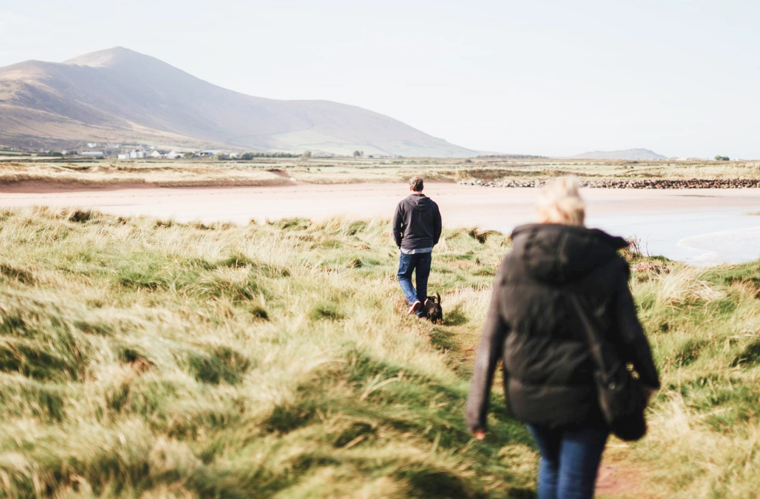 A person in a dark jacket walks through windswept coastal grassland toward another hiker in the distance, with a calm bay and mountain backdrop under overcast skies.