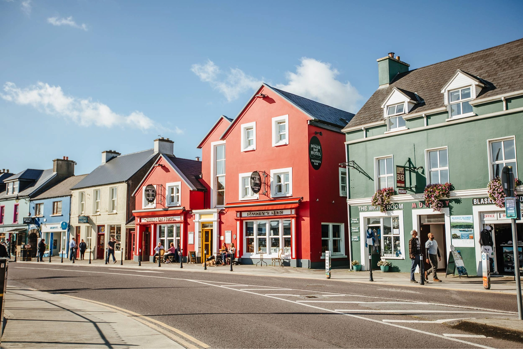 Colorful shop fronts line a pedestrian street in Dingle, including a prominent red building with shop windows and residential spaces above, alongside pastel-colored neighboring buildings under a partly cloudy sky.