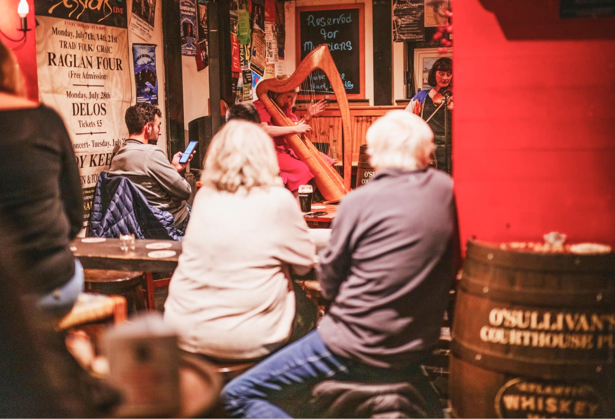 Visitors seated at tables in a traditional Irish pub with red walls, watching a musician play a harp on stage. Posters and whiskey barrel decorations line the walls of this intimate venue in Dingle.