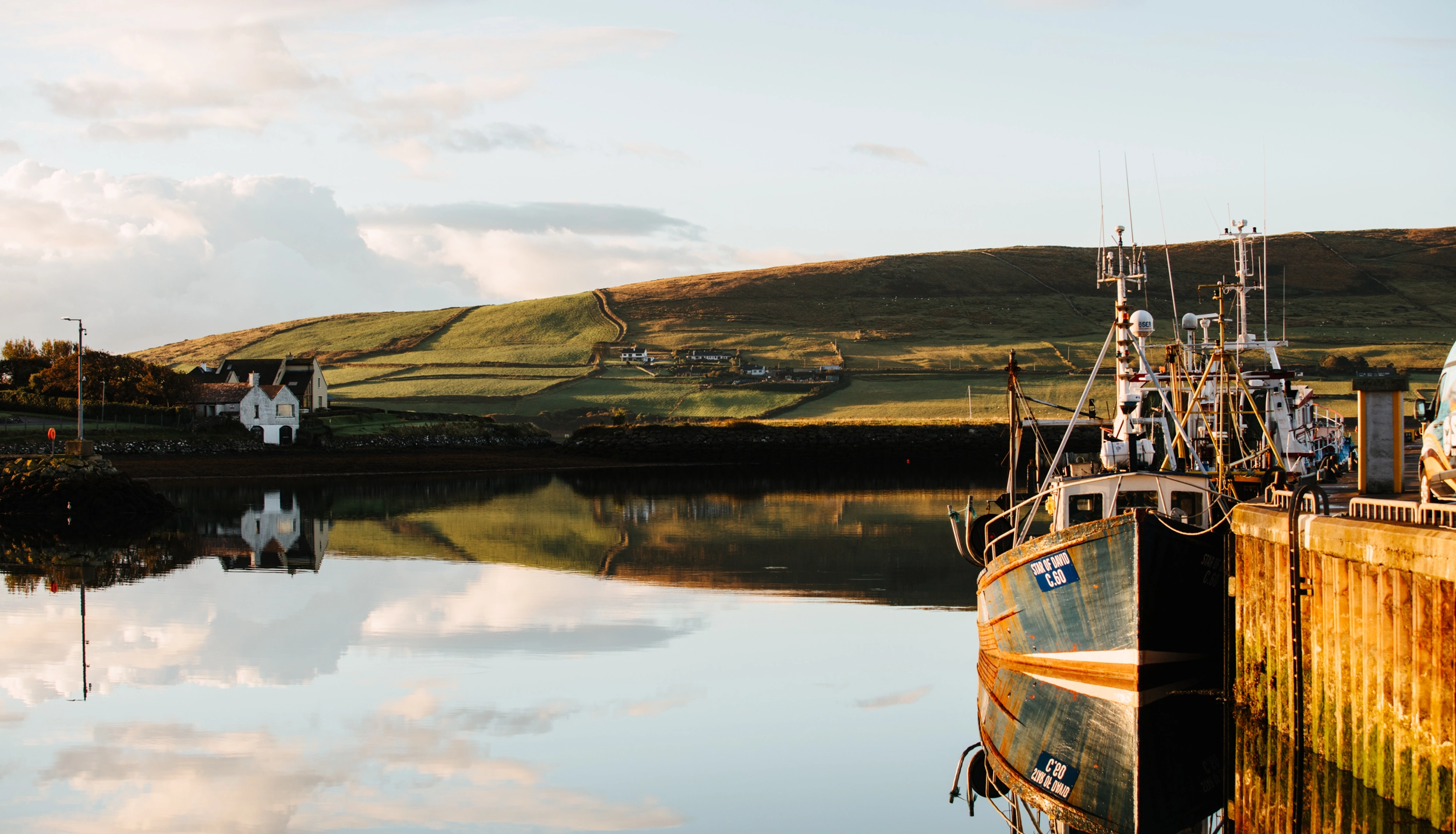 A fishing vessel with yellow and blue markings is moored at a wooden dock in a calm harbor, with rolling green hills and white buildings reflected in the still water under a partly cloudy sky.