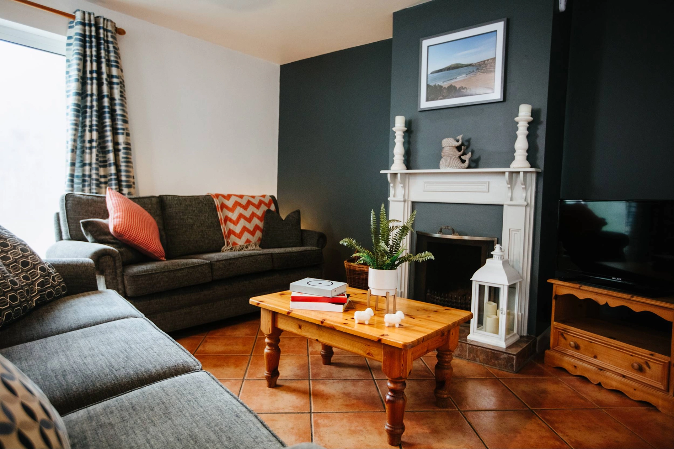 A cozy living room with dark teal accent walls, a brown sectional sofa with orange and patterned cushions, and a wooden coffee table on hardwood floors. A white fireplace mantel with decorative items sits against the feature wall, with a framed landscape photo above it and a TV to the right.