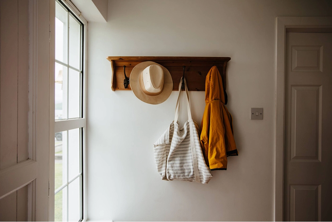 A wooden wall-mounted coat rack with hooks holds a tan straw hat, a striped cream and beige canvas tote bag, and a mustard yellow cardigan in a bright hallway near a window and doorway.