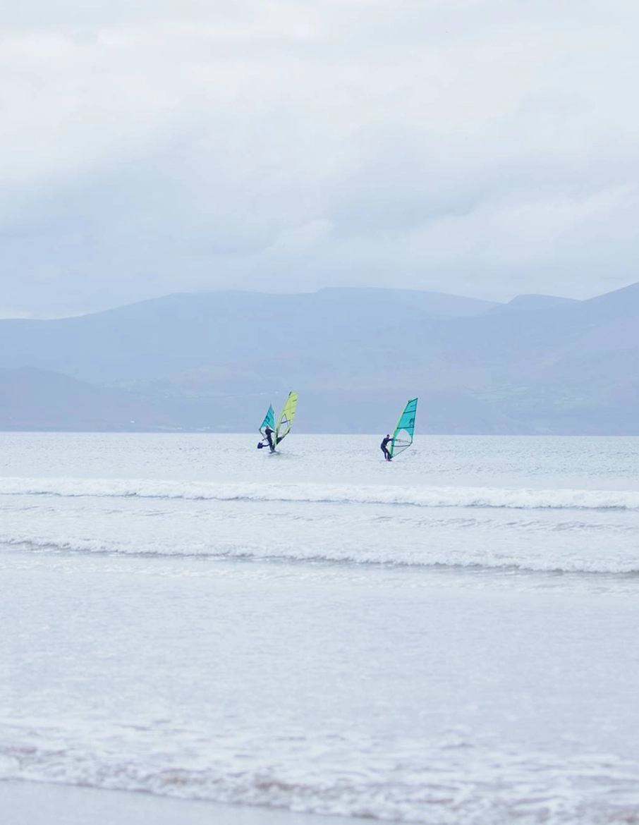 Two windsurfers with colorful sails navigate the calm waters of Dingle Harbour on an overcast day, with mountains visible across the bay in the background.