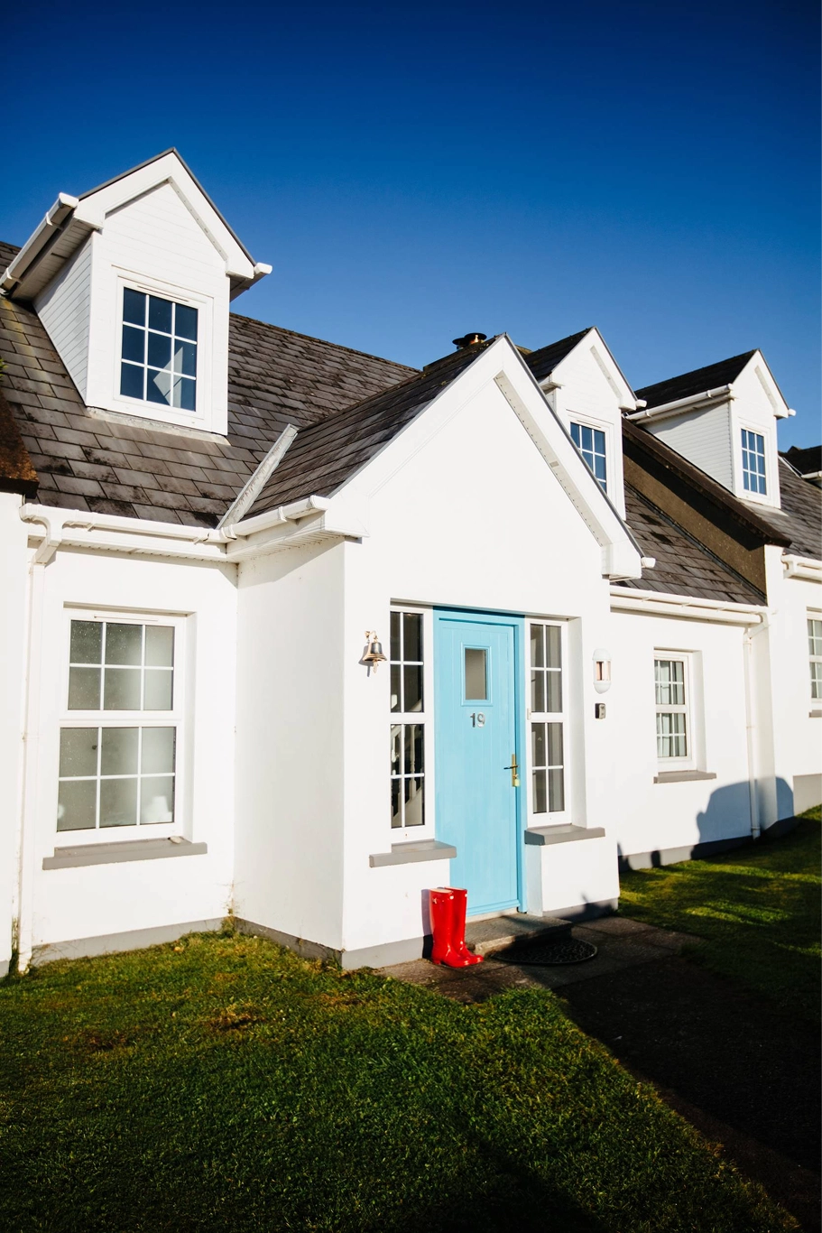 A white cottage with a bright turquoise blue front door, multiple dormer windows, and manicured green lawn under a clear blue sky at Dingle Harbour Cottages.