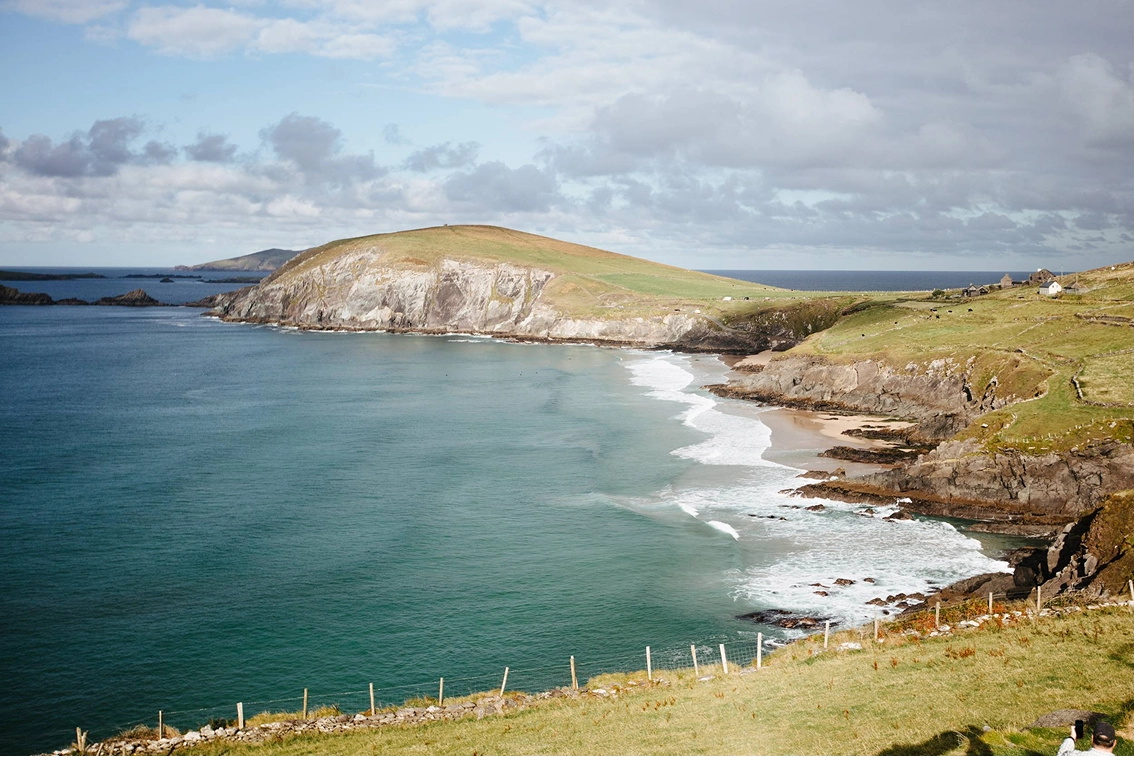 A dramatic coastal headland with white chalk cliffs rises above turquoise waters and a sandy beach, with rolling green hills and farmland stretching across the peninsula under a partly cloudy sky.