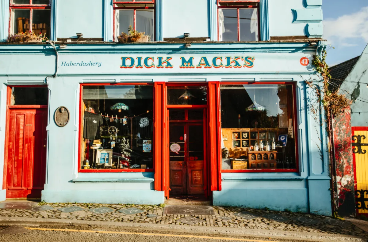 Dick Mack's traditional pub with light blue exterior walls, red-framed windows and door, located on a cobblestone street in Dingle. The storefront displays 