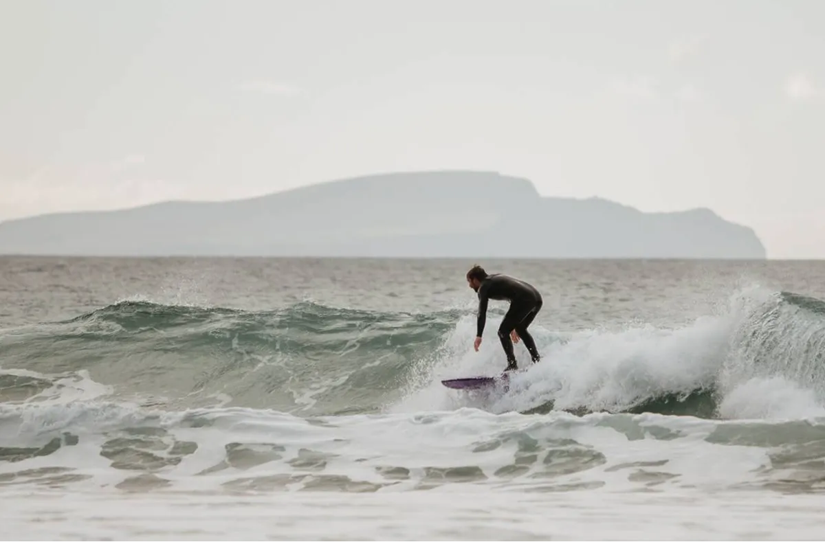 A surfer in a wetsuit rides a wave on a surfboard at a beach, with Table Mountain visible across the water in the misty distance.