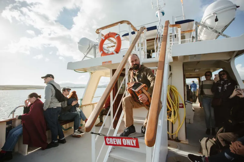 A crew member assists passengers boarding a tour boat at Dingle Harbour, with visitors climbing wooden stairs to enter the vessel while others wait on the dock. The boat is equipped with safety equipment including a life ring and satellite equipment visible on its white exterior.