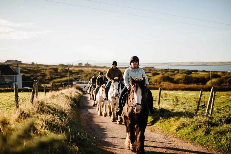 Two riders on horseback travel along a rural dirt path lined with wooden fences, with golden grassland and a distant blue bay visible under an overcast sky.