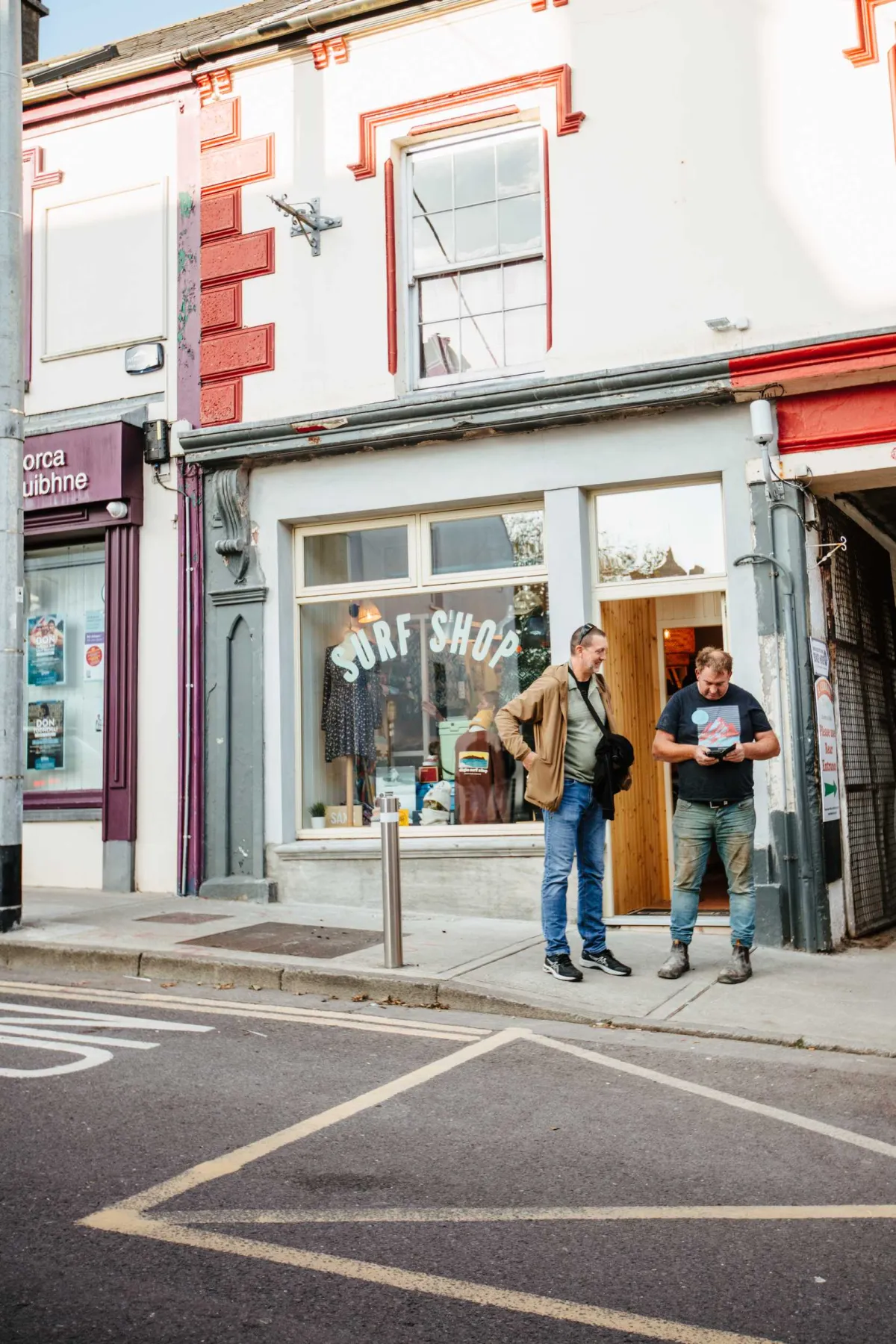 Two people stand in the doorway of a surf shop with large storefront windows on a narrow street. The shop displays surfboards and equipment behind the glass, with orange and white architectural details above. Street markings are visible on the pavement in front of the storefront.