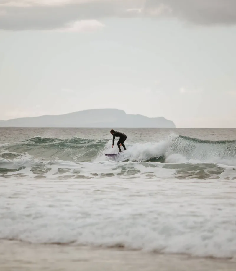 A surfer in a wetsuit rides a wave on his surfboard in the Atlantic Ocean near Dingle, Ireland, with a flat-topped mountain visible across the water under an overcast sky.
