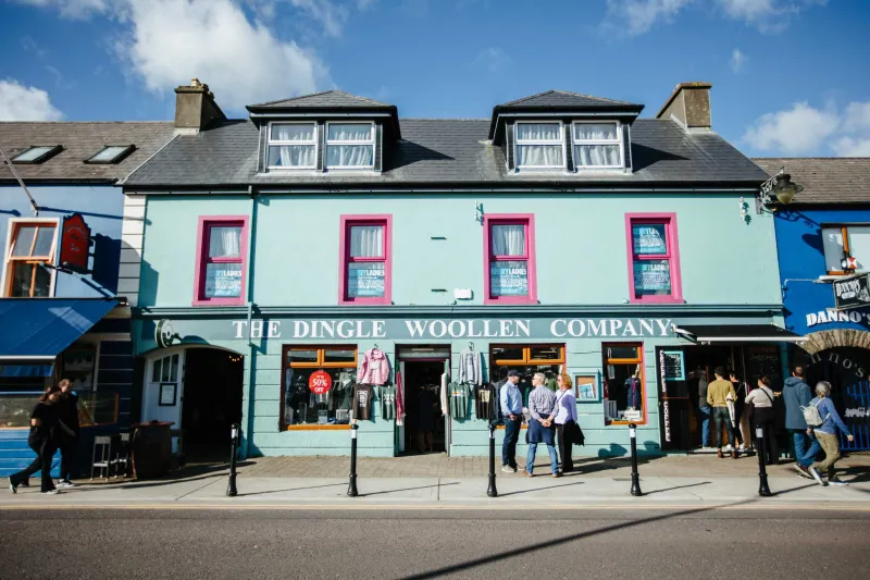 A turquoise-colored storefront of the Dingle Woollen Company with bright pink window frames and doors on a pedestrian street in Dingle, Ireland, with tourists browsing outside and traditional stone buildings with dormer windows above.