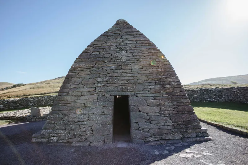 A stone beehive-shaped oratory with a narrow doorway stands on a grassy hillside under clear skies. The structure, built from stacked slate-grey stones, features a distinctive conical roof and is surrounded by rolling green fields and distant hills. This ancient Irish stone building exemplifies early Christian architecture.