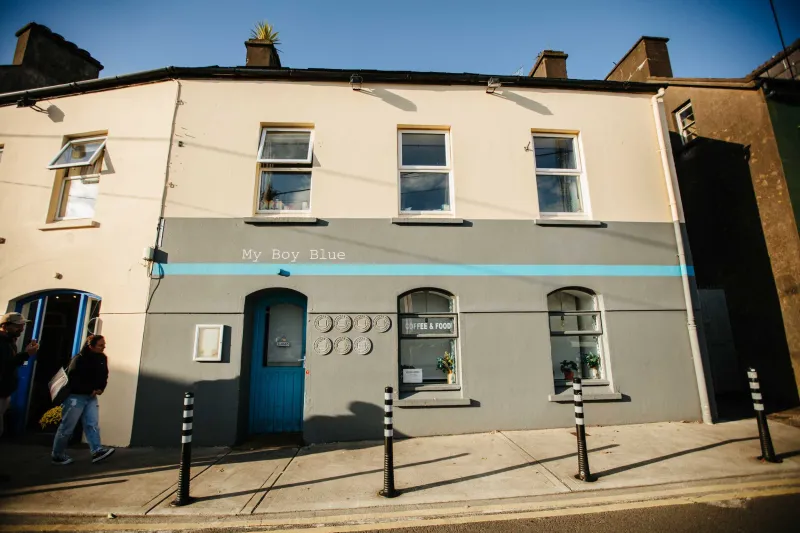 A two-story storefront with cream-colored upper walls and gray lower walls featuring a turquoise stripe. The ground floor has a blue door, phone keypad, and window displays for 