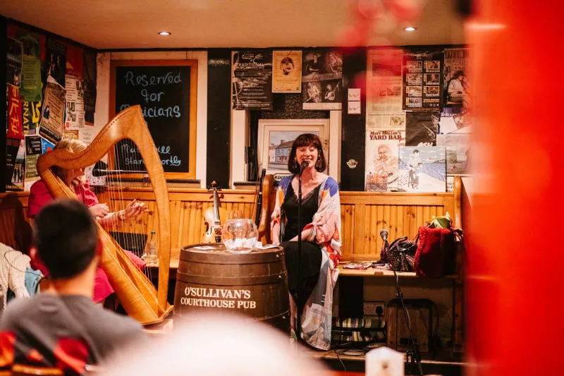 A musician plays harp while a woman sings at O'Sullivan's Courthouse Pub in Dingle, with warm amber lighting and traditional pub décor visible in the background.