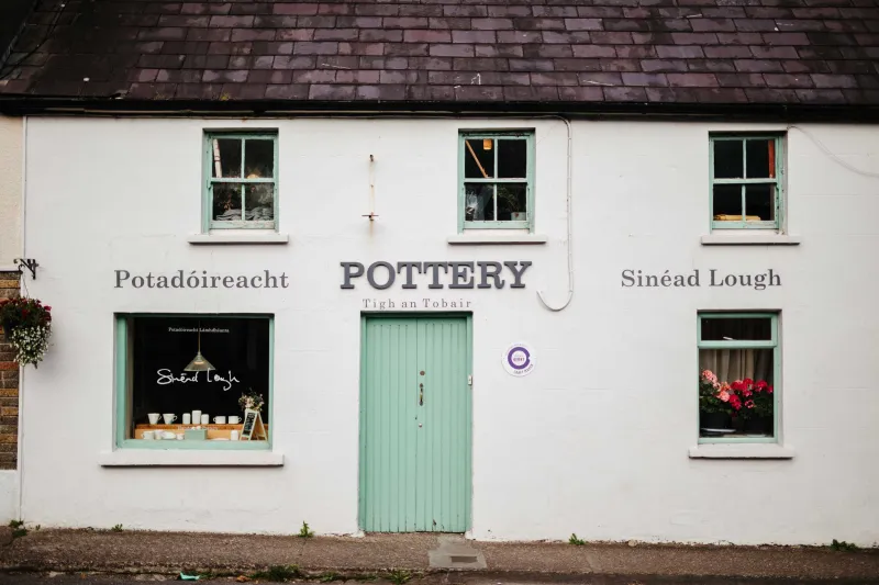 A white-walled pottery shop with a mint green door and three windows set in a dark slate roof. The storefront displays bilingual signage reading 