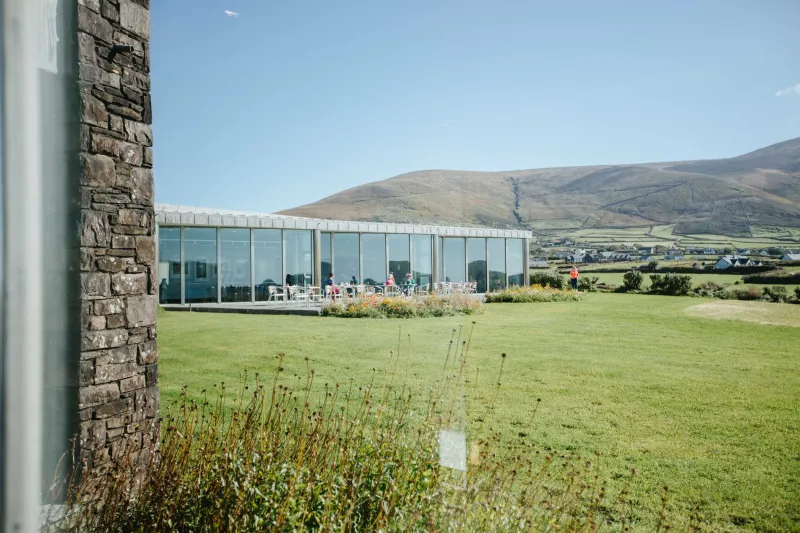 A modern glass-walled building with stone pillars sits on a grassy hillside overlooking a green valley with mountains in the background under clear blue sky.