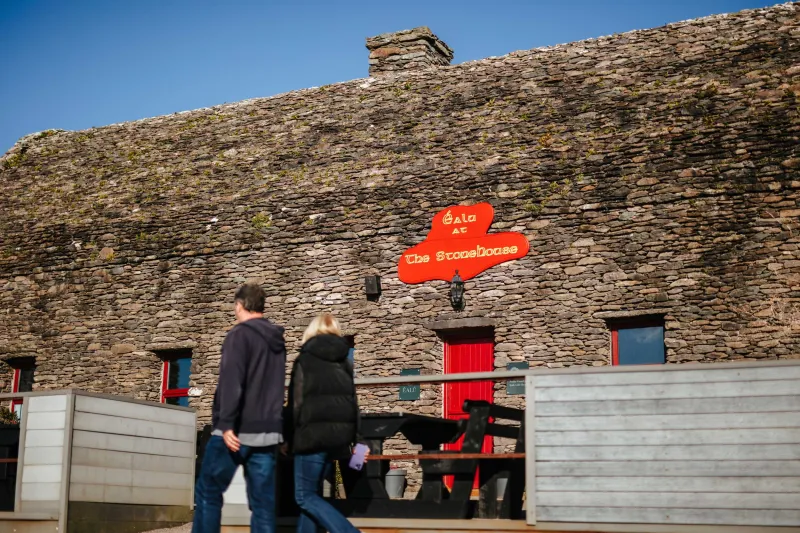 Two visitors stand on a wooden platform viewing a striking red directional sign mounted on a stone cottage wall. The rustic stone building features small windows and a red door, with the arrow sign pointing to various local destinations.