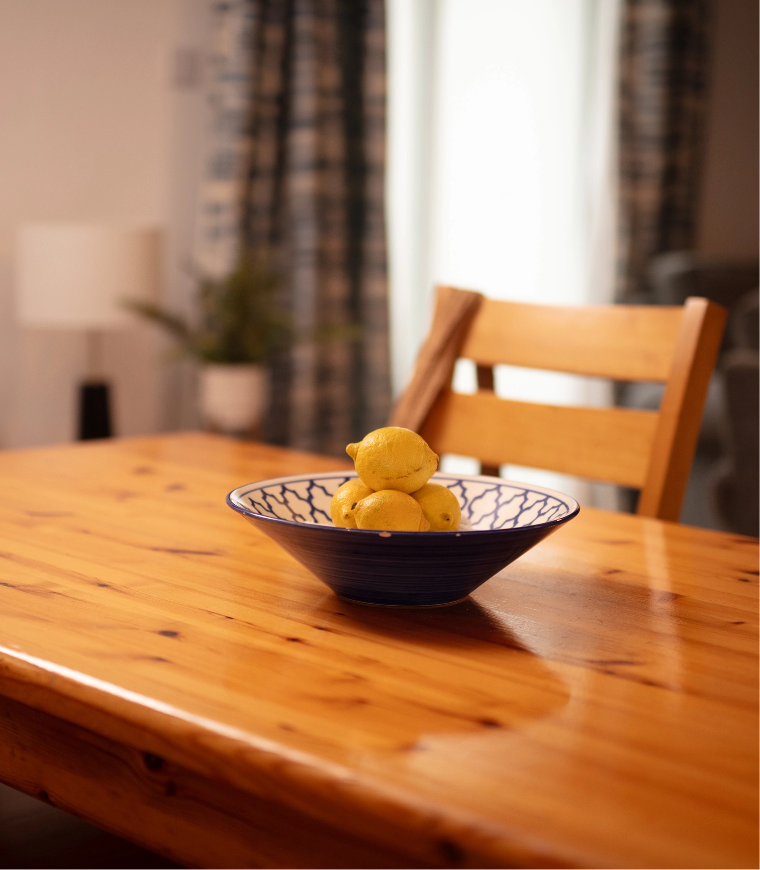 A wooden dining table with a blue decorative bowl filled with yellow lemons, wooden chairs, and a bright window with patterned curtains in the background.