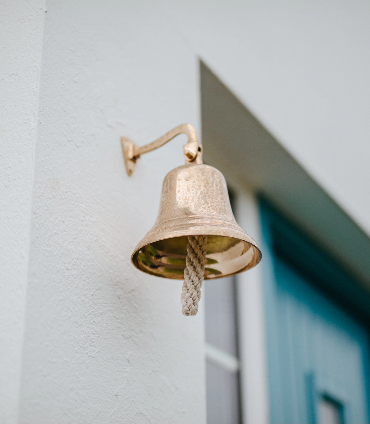 A brass bell mounted on a white wall with a rope pull, casting a shadow in bright sunlight near a blue-painted doorway of a cottage.