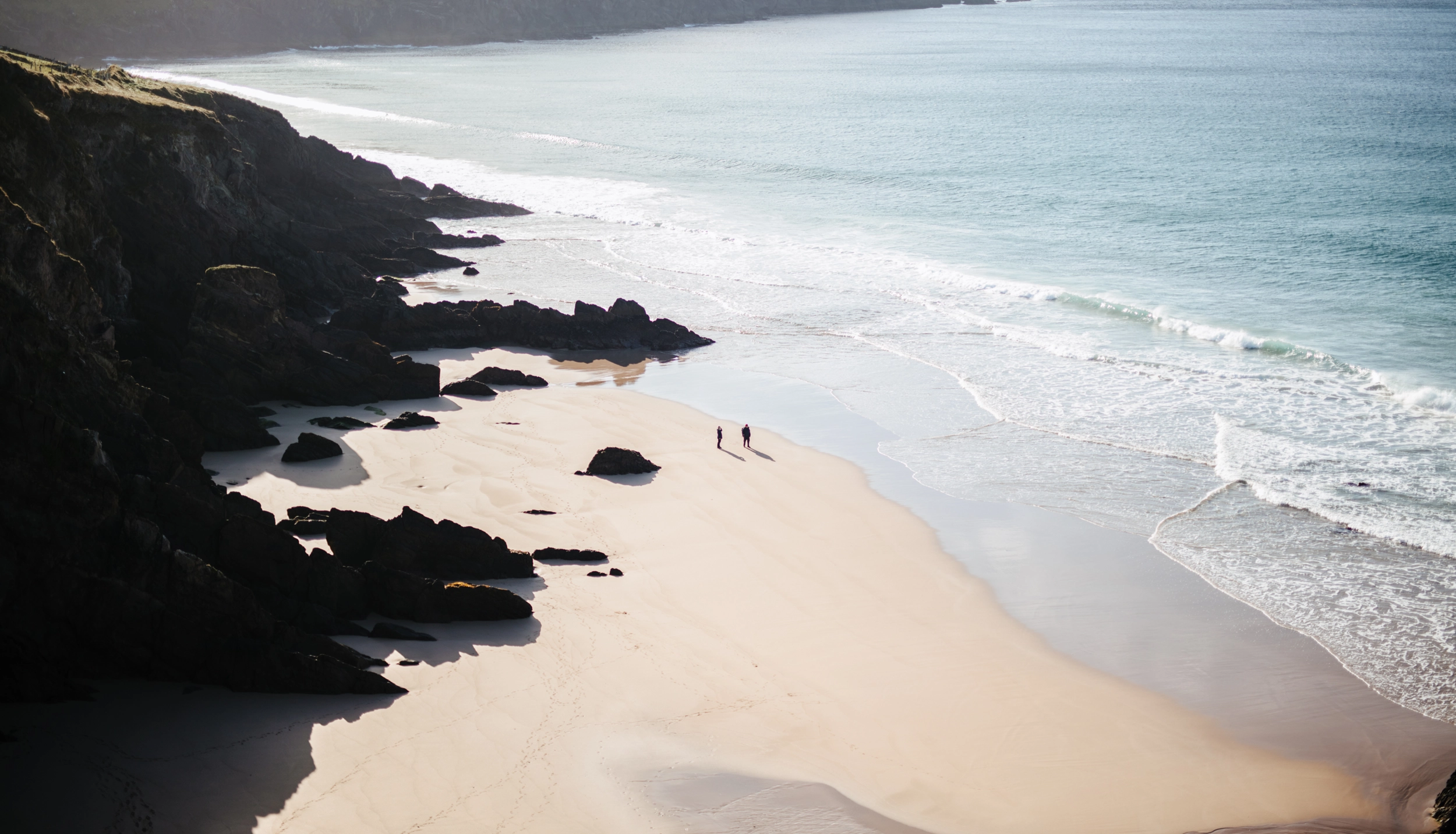 A pristine sandy beach curves along dramatic dark rocky cliffs, with turquoise waters and white-capped waves rolling onto the shore. Two small figures stand on the sand near the water's edge. The secluded cove, framed by rugged black rock formations, exemplifies the scenic coastal landscape near Dingle Harbour.