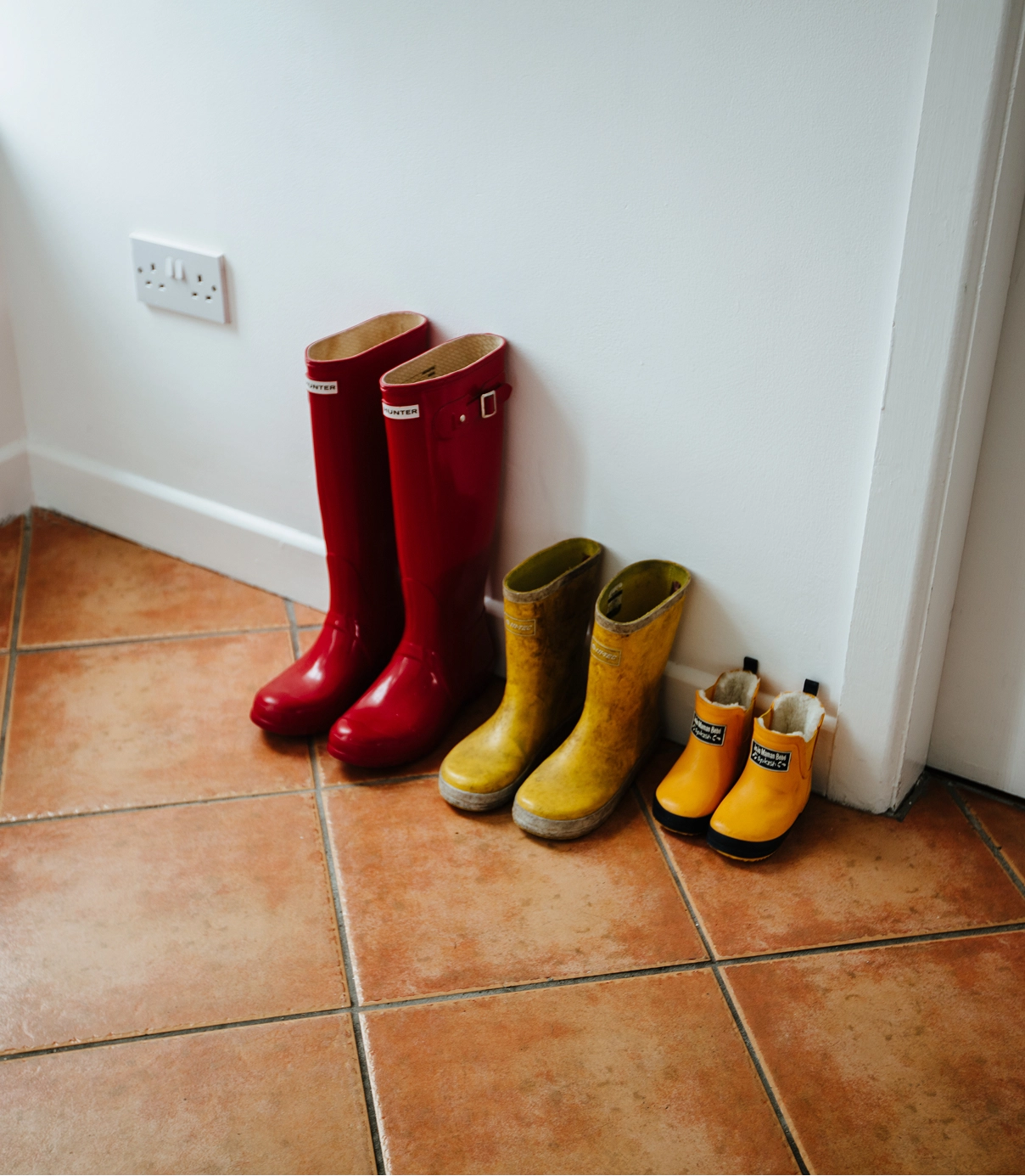 A collection of wellington boots in varying sizes arranged on a terracotta tile floor against a white wall, including red adult boots, yellow boots, and smaller children's boots in orange and yellow.