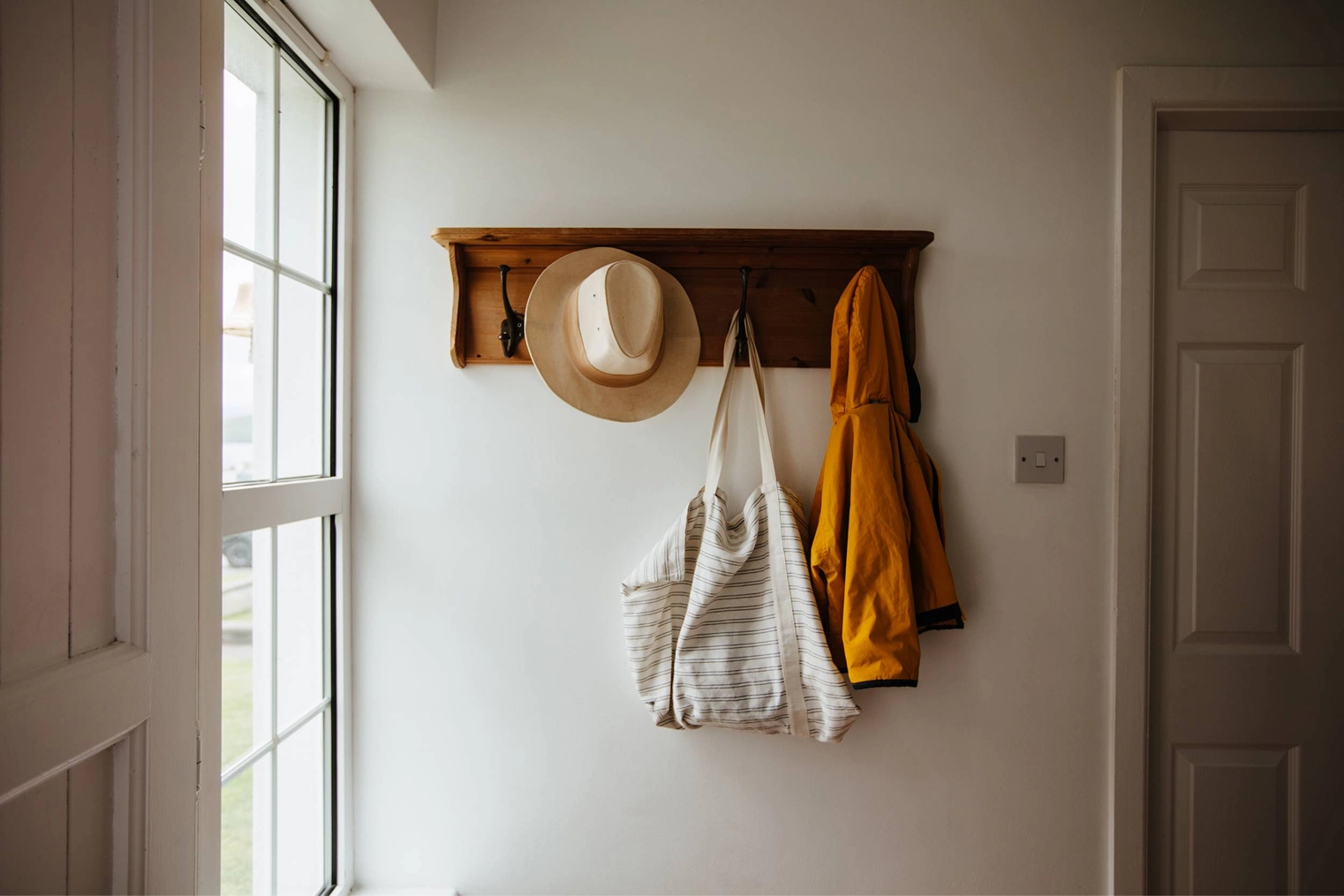 A wooden wall-mounted coat rack holds a tan straw hat, striped linen tote bag, and mustard-yellow cardigan beside a bright window and doorway in a cottage entryway.