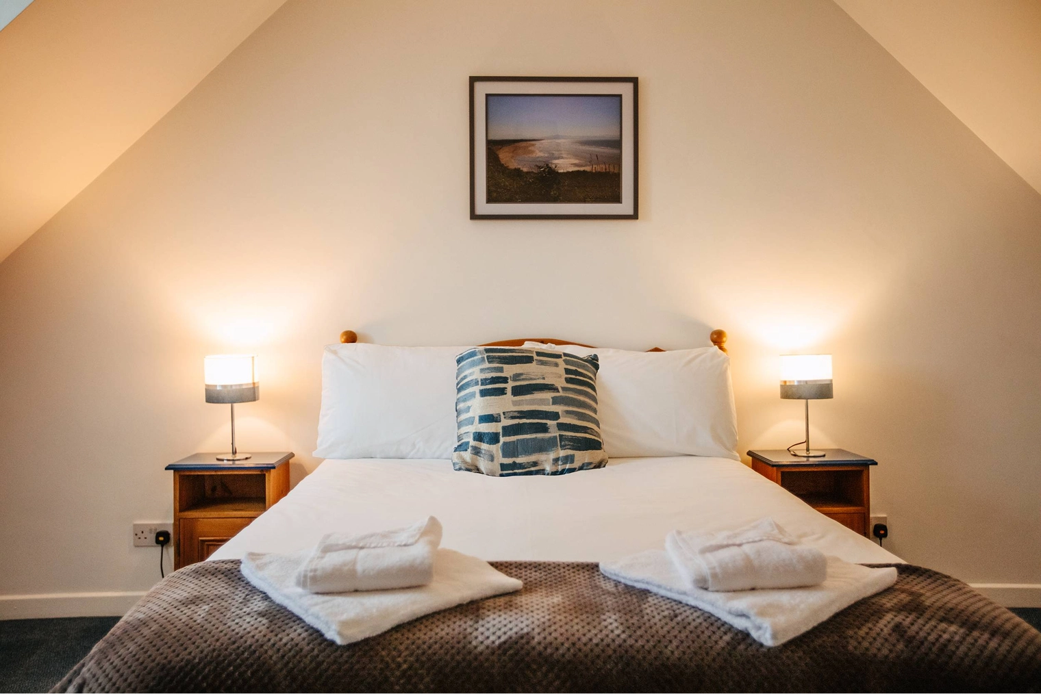 A neatly made double bed with white pillows and a blue patterned cushion in a bright attic bedroom with sloped ceilings, flanked by wooden nightstands with warm lamps, and a framed landscape photograph on the wall above.