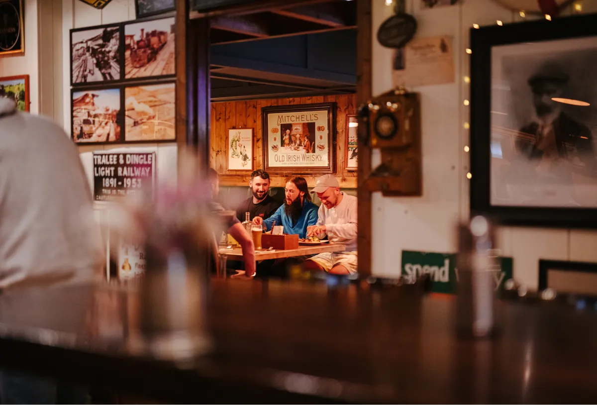 Two people sit at a table inside a warmly-lit cottage, with framed photographs and decorative elements visible on the walls behind them. The scene is viewed through a window or glass barrier with blurred objects in the foreground. The cozy interior features wooden beams and ambient lighting creating a homey atmosphere.