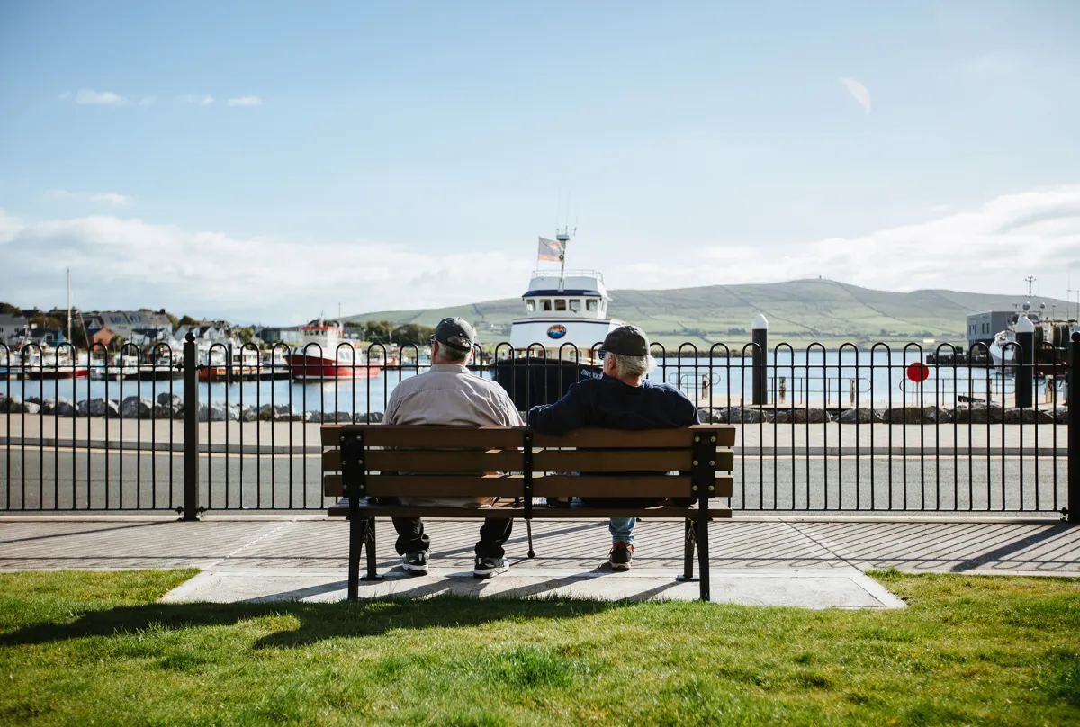 Two people sit on a wooden bench facing Dingle Harbour, with a blue and white ferry boat and colorful waterfront buildings visible across the water, mountains in the distance under a partly cloudy sky.