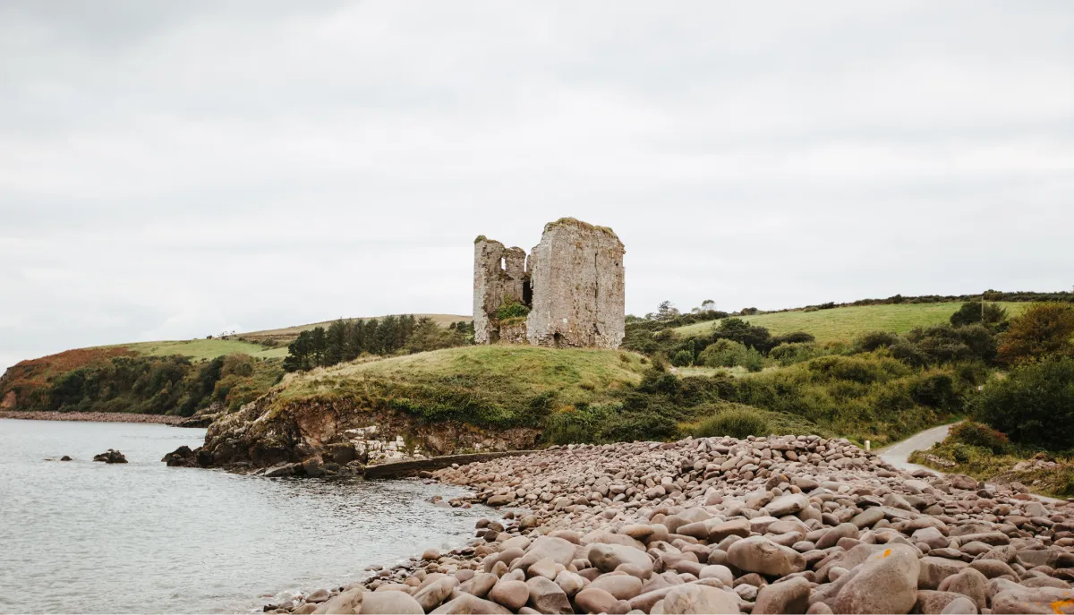 Ruins of a stone tower stand on a grassy headland overlooking a rocky beach and calm harbor water under an overcast sky in Dingle, Ireland.
