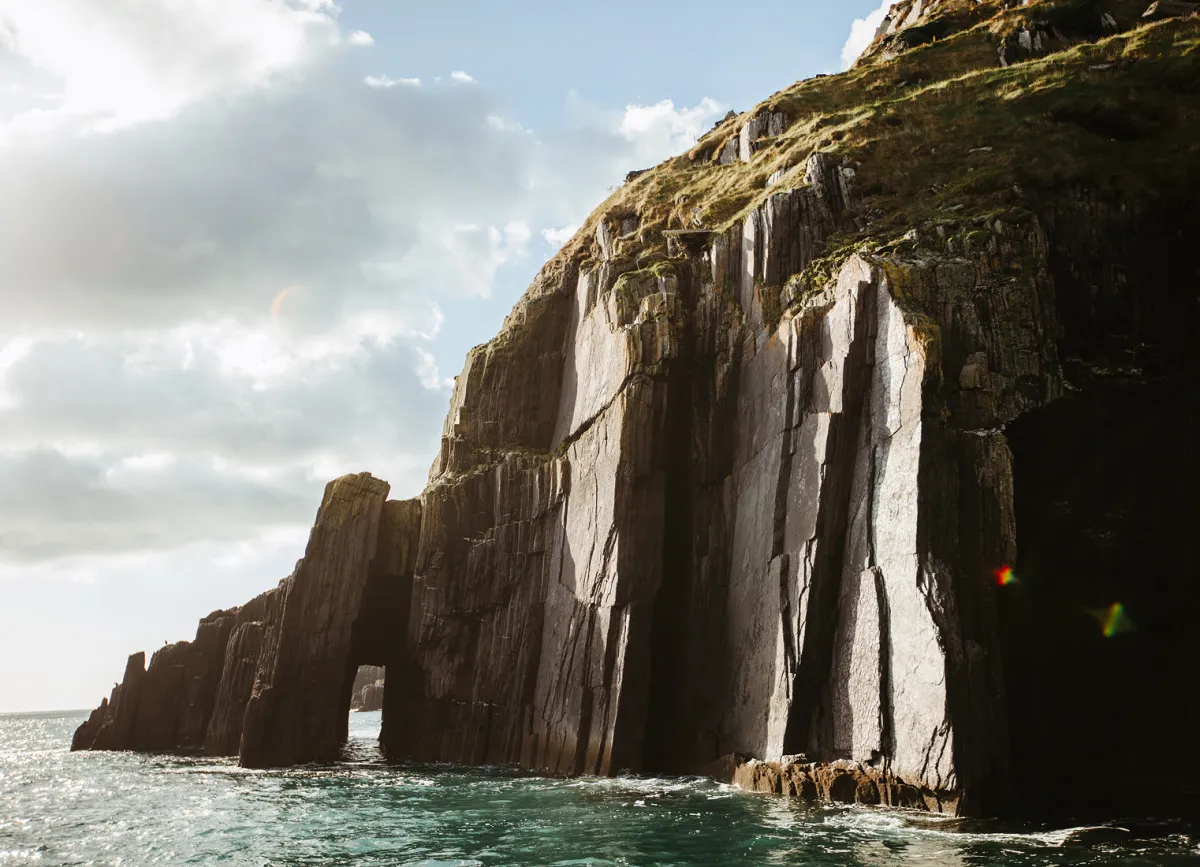 Dramatic basalt sea cliffs with distinctive columnar rock formations rise from turquoise waters, their dark stone faces streaked with white mineral deposits and lichen.