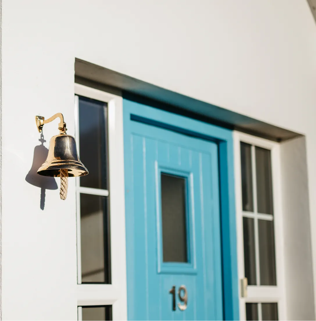 A brass ship's bell hangs from a bracket mounted on a white-painted wall beside a bright turquoise blue cottage door numbered 19, with white-trimmed windows flanking the entrance.