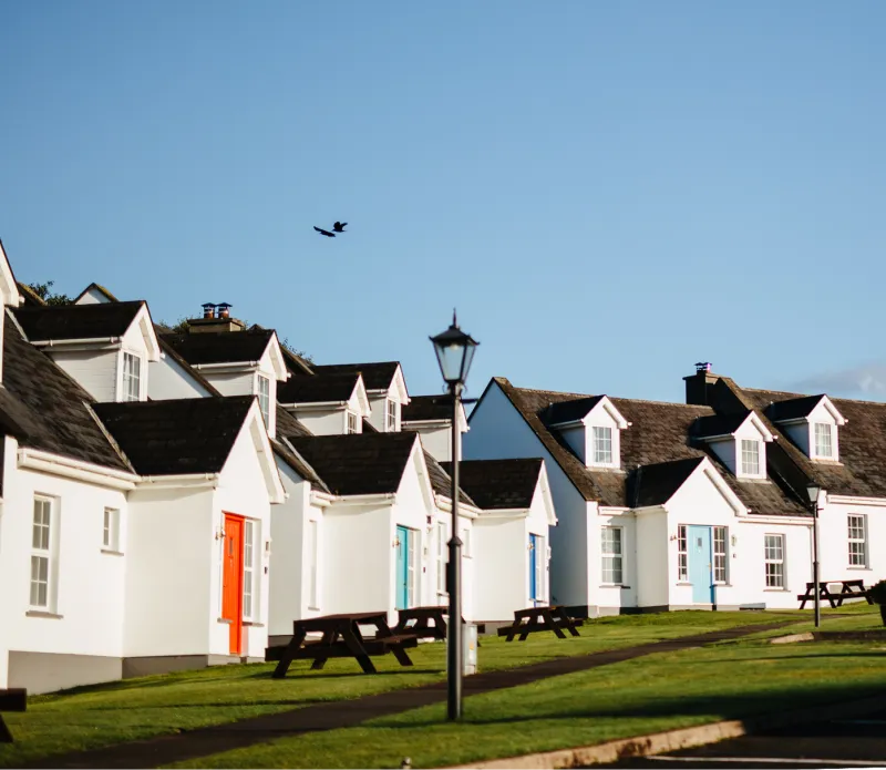 A row of white-painted cottage accommodations with dark roofs and colorful front doors sits on a grassy waterfront lawn under clear blue sky, with a traditional street lamp and picnic tables visible in front of the buildings.